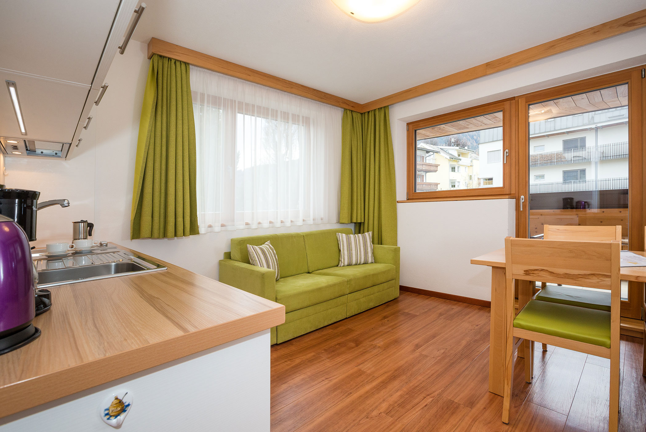 Cucina e zona pranzo luminose con divano verde, tavolo in legno e ampie finestre con tende verdi. - Residence Aichner