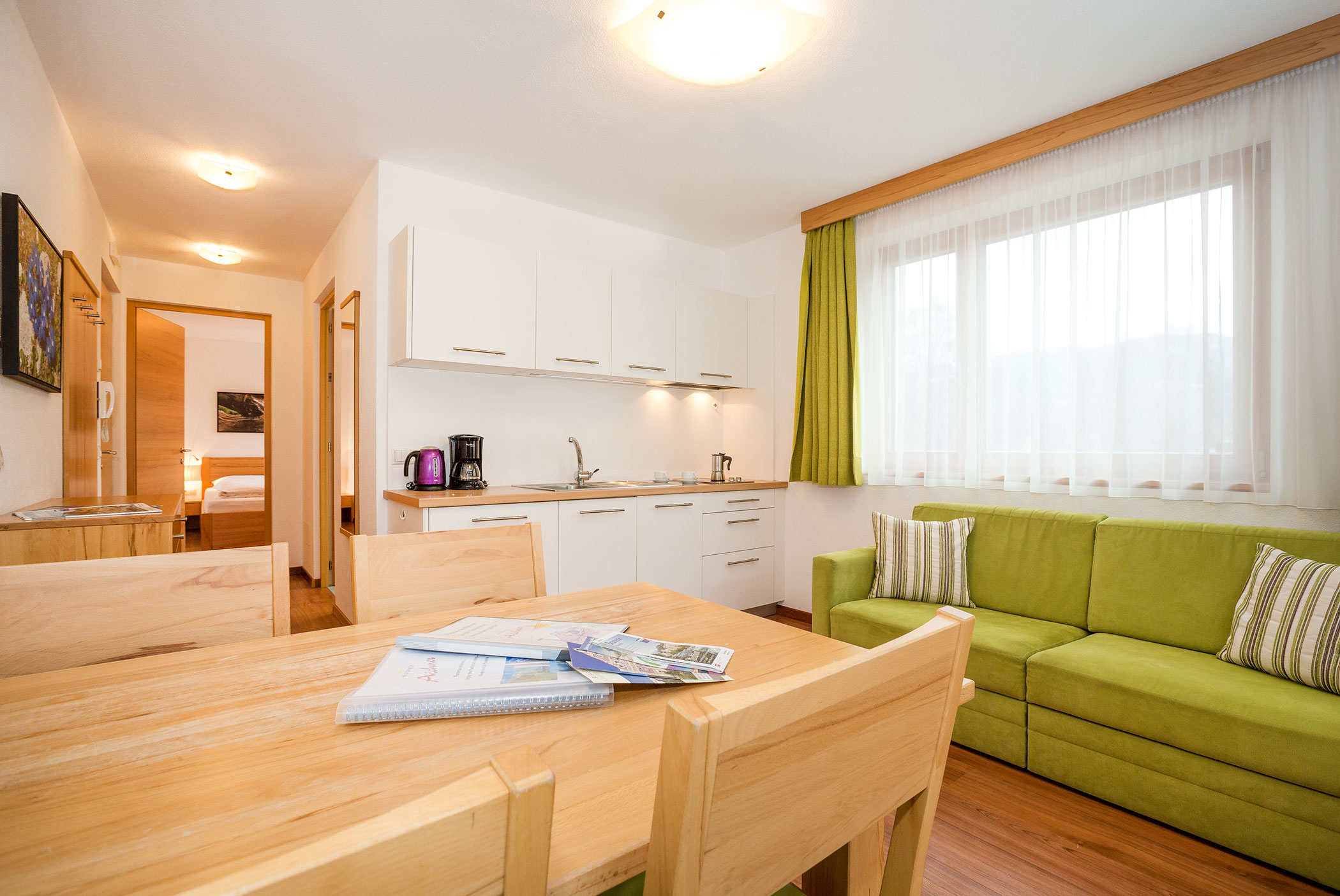 Cucina luminosa e zona giorno con un divano verde, un tavolo in legno e una grande finestra con tende. - Residence Aichner