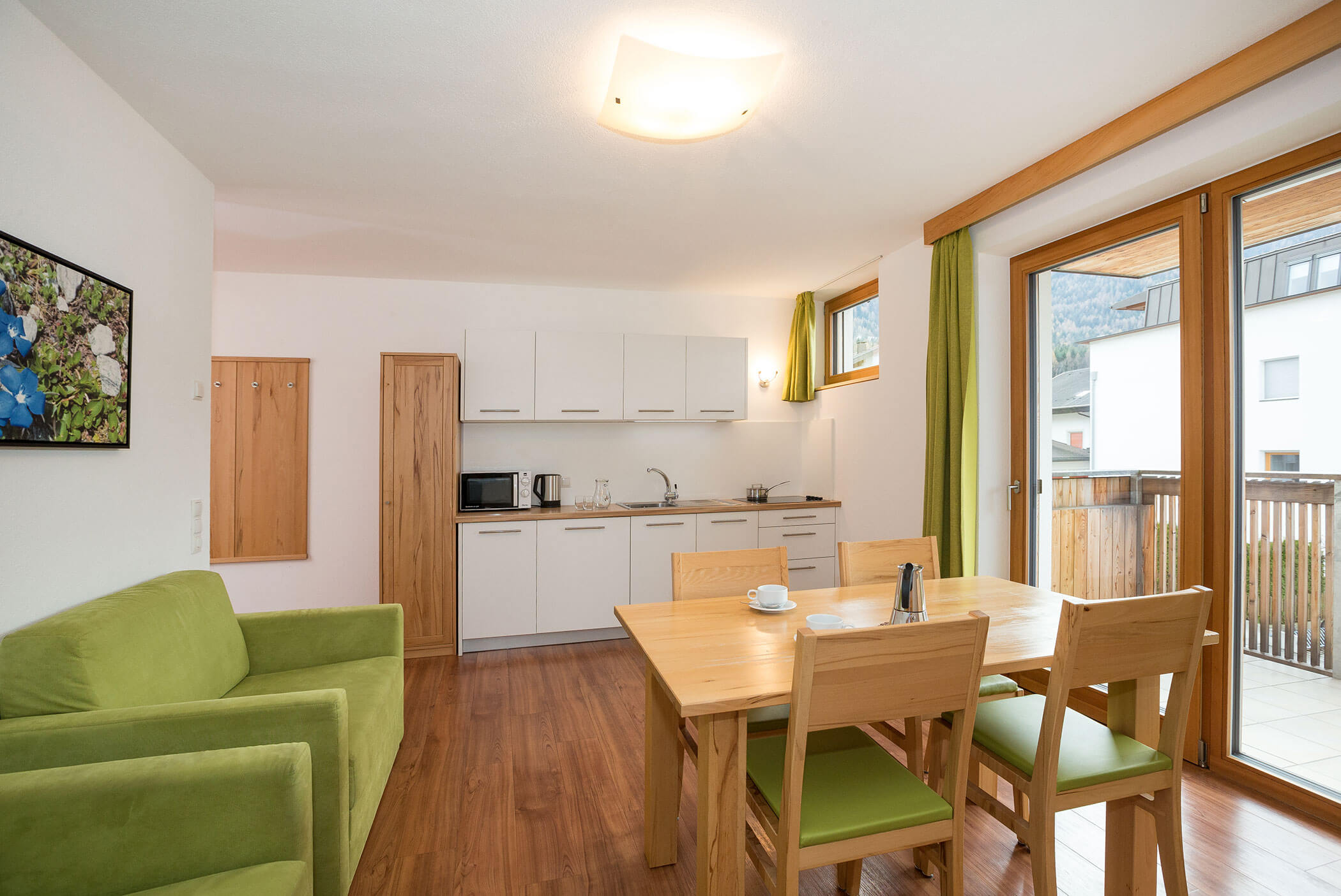 Cucina moderna e zona pranzo con divano verde, tavolo in legno e ampie finestre che lasciano entrare la luce naturale. - Residence Aichner
