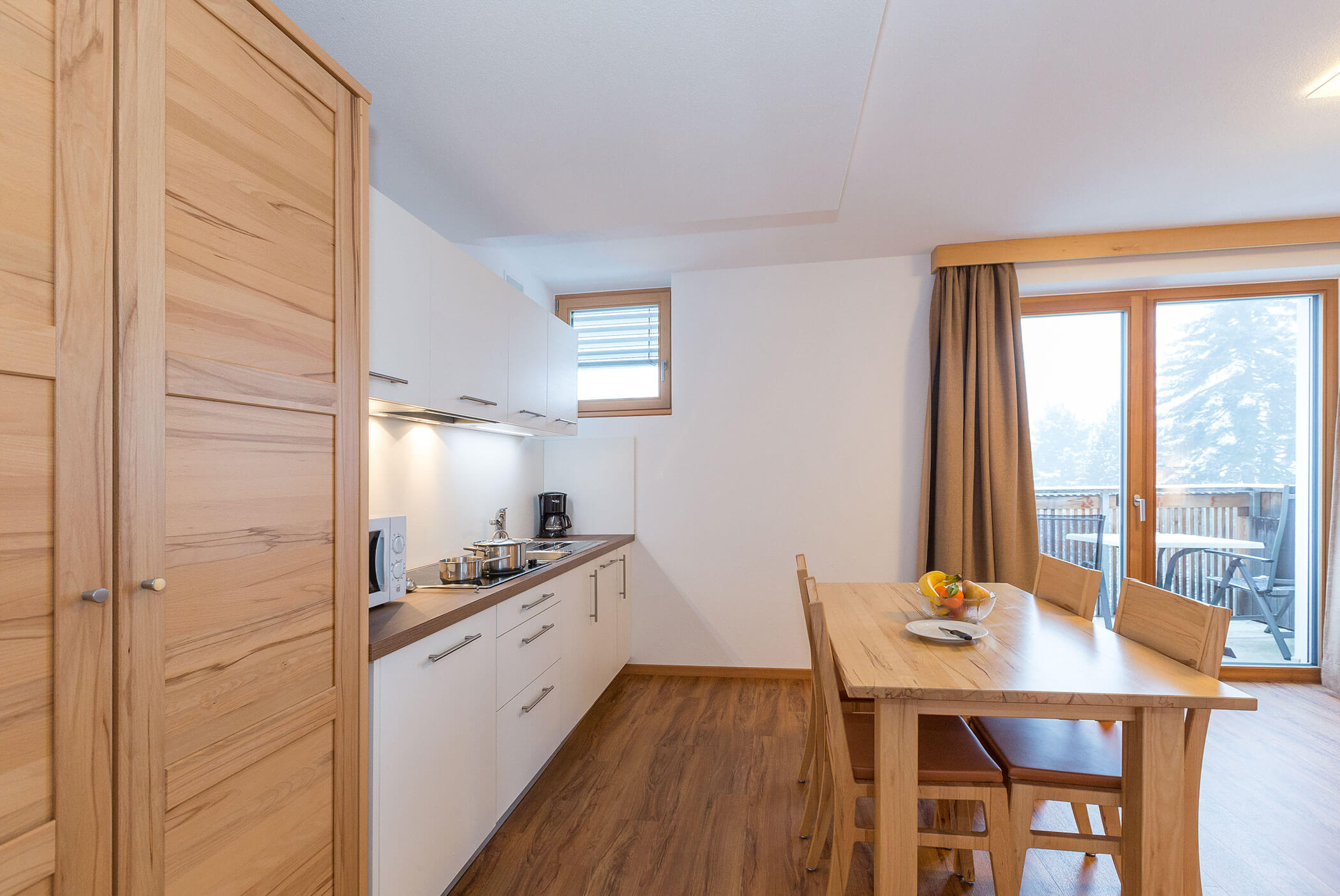 Cucina moderna con mobili bianchi, tavolo e sedie in legno e vista sul balcone grazie alle ampie finestre. - Residence Aichner
