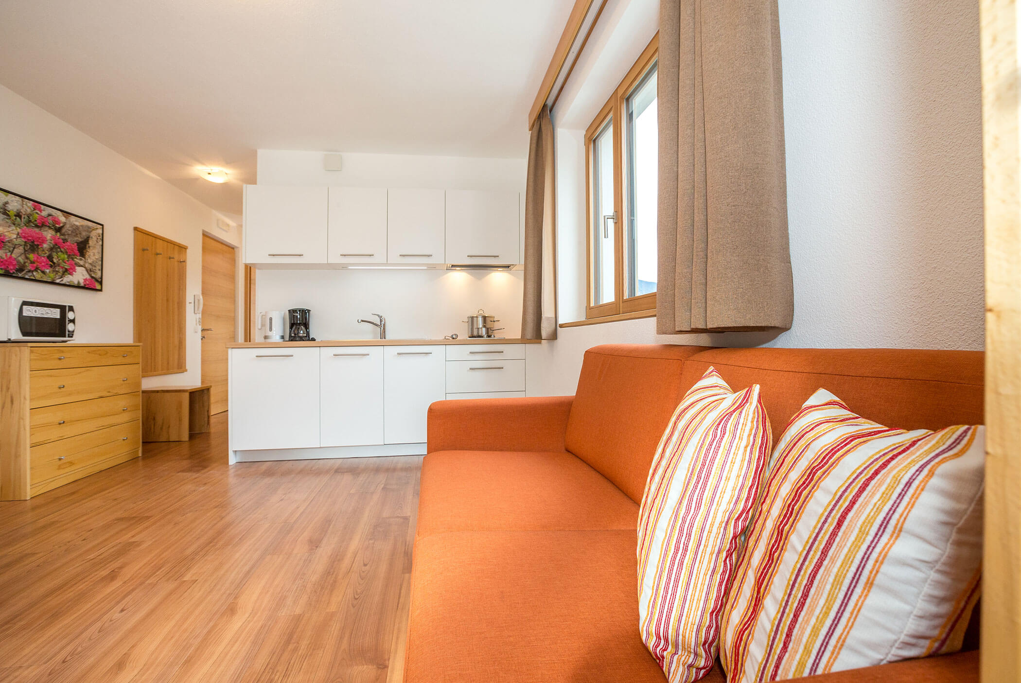 Moderno monolocale con divano arancione, cuscini a righe, pavimenti in legno e angolo cottura bianco con elettrodomestici. - Residence Aichner