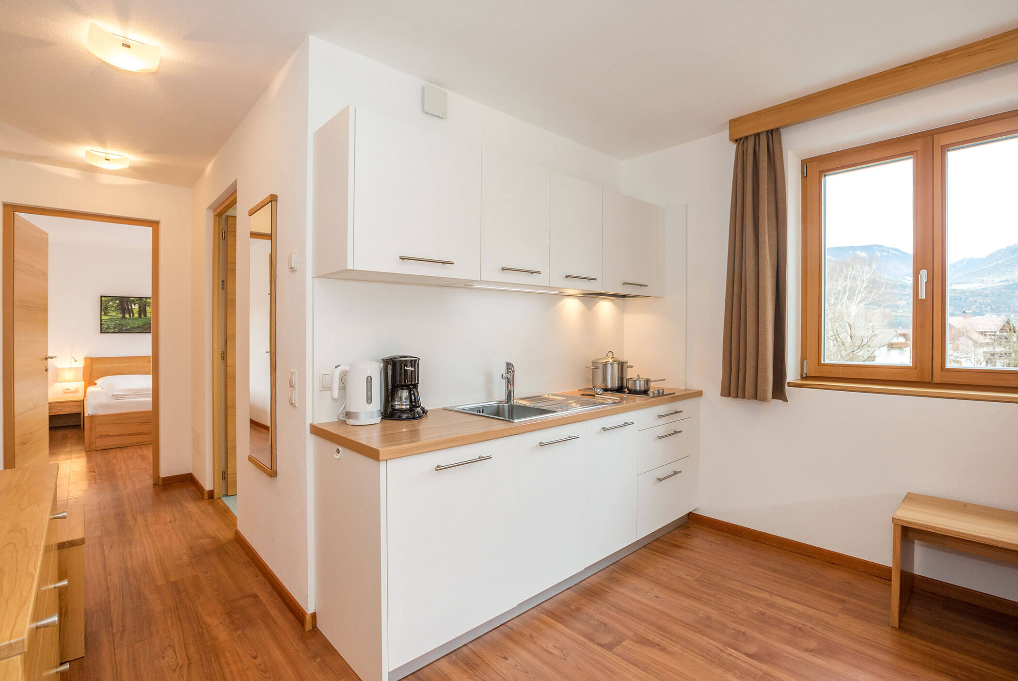 Cucina moderna con mobili bianchi, piani di lavoro in legno e una finestra; dalla porta aperta si intravede la camera da letto. - Residence Aichner