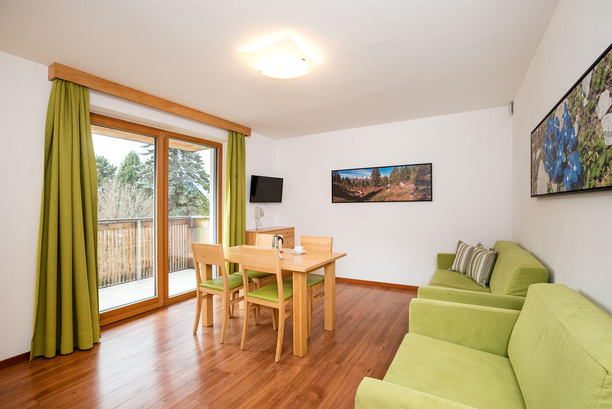 Sala da pranzo luminosa con divani verdi, tavolo in legno, sedie, quadri alle pareti, TV e portefinestre con tende verdi. - Residence Aichner