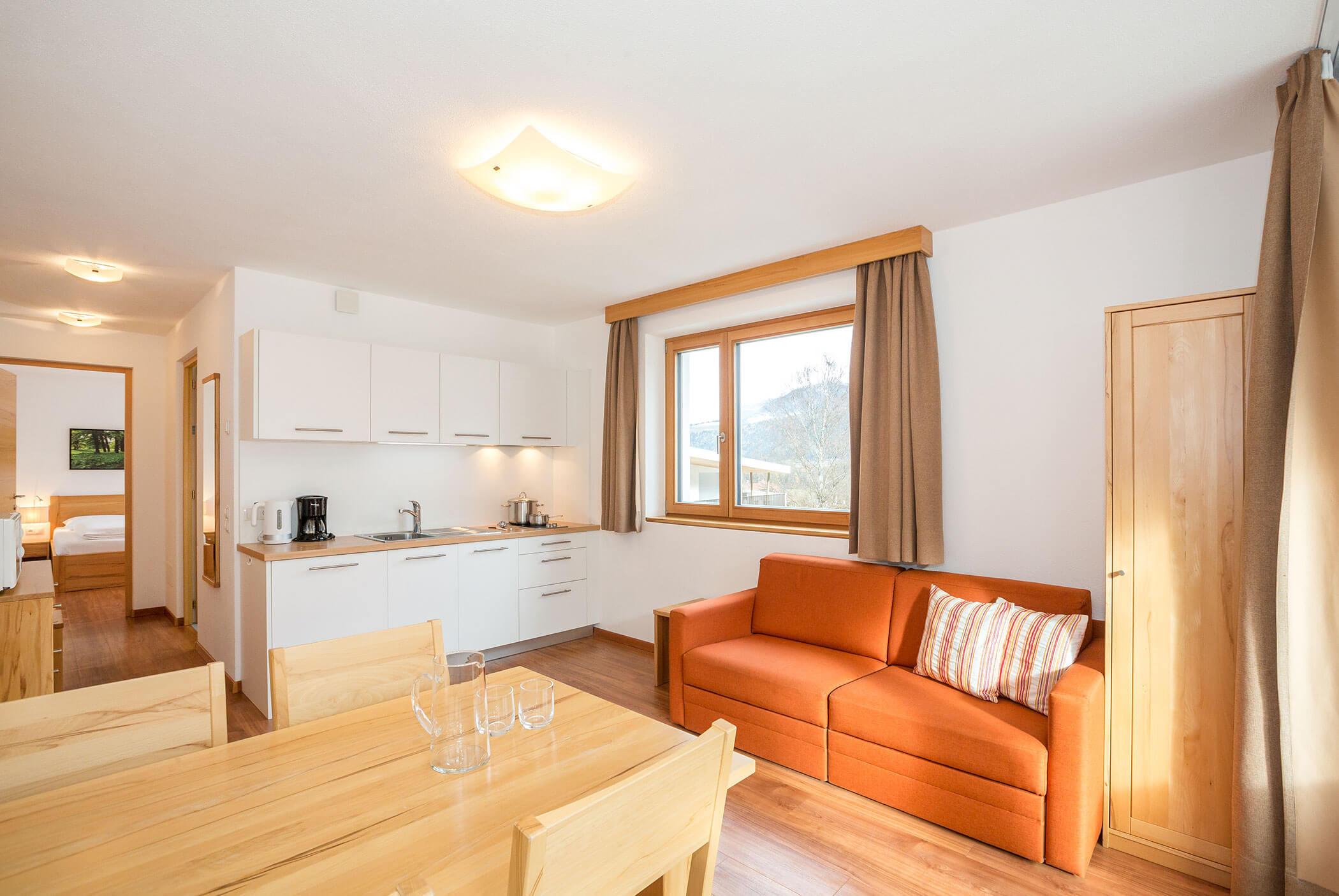 Luminosa zona cucina-soggiorno con divano arancione, tavolo da pranzo e ampia finestra; in fondo si intravede la camera da letto. - Residence Aichner